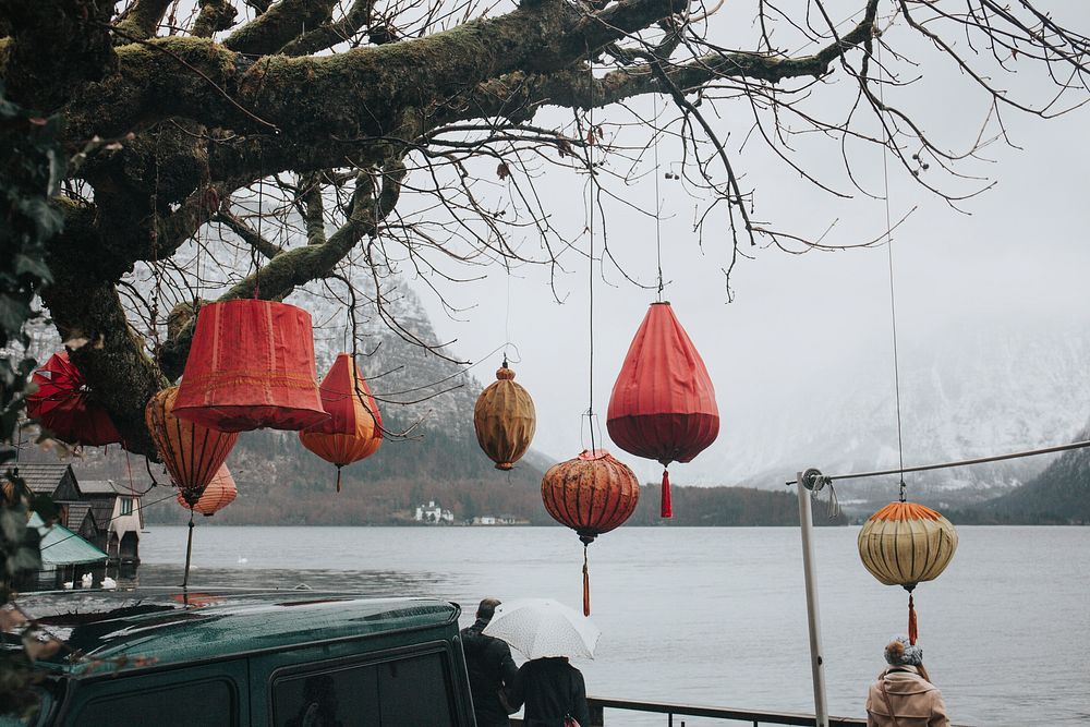 Colorful red paper lamps hang from a tree overlooking the water and a winter landscape. Original public domain image from…