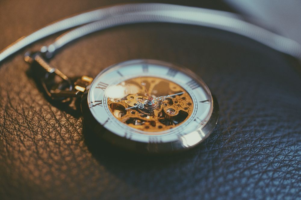 An intricate pocket watch showing roman numerals and a complex gear system behind the hands resting on a leather piece of…