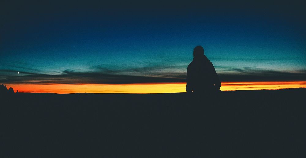 Silhouette person standing in foreground of a black, orange and blue sunrise-or-sunset horizon in Montana. Original public…