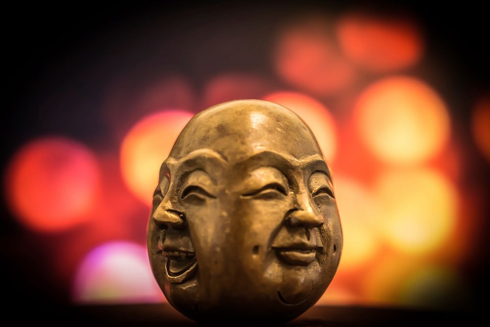 A brass figurine depicting two smiling faces of Buddha, with blurry colored lights in the background. Original public domain…