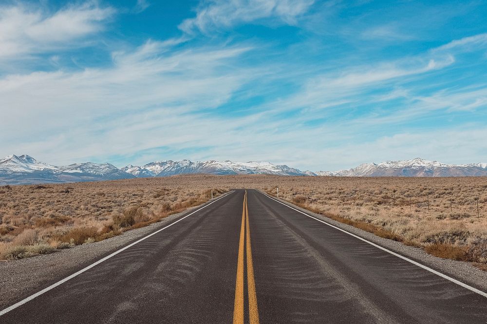 A long empty asphalt road through dry plains with snowy mountains on the horizon. Original public domain image from…