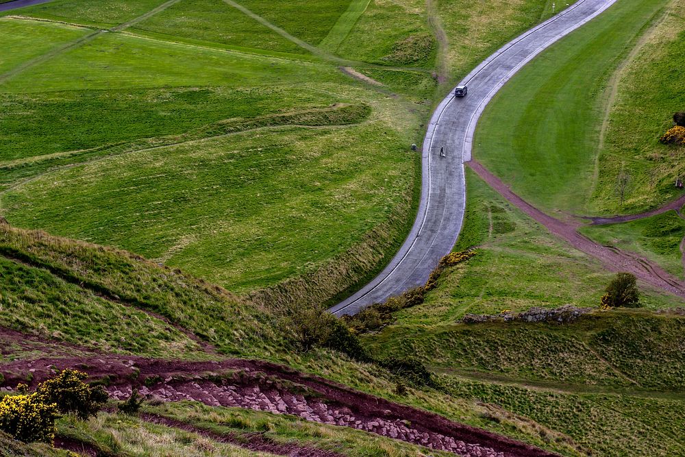 Drone shot of car driving on countryside road surrounded by green fields in Edinburgh, Scotland. Original public domain…