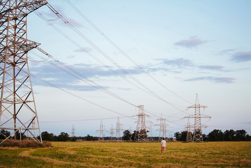 Man in a white shirt and red shorts standing in a field gazing at the pylons and power lines running between them under a…