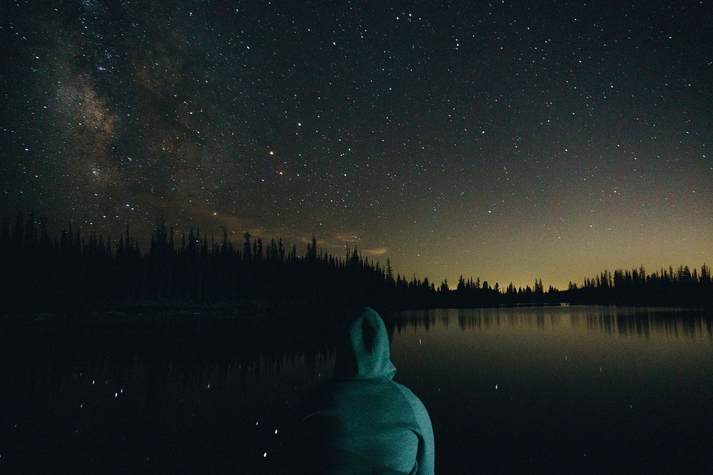 A hooded person sitting near a lake under a starry sky. Original public domain image from Wikimedia Commons