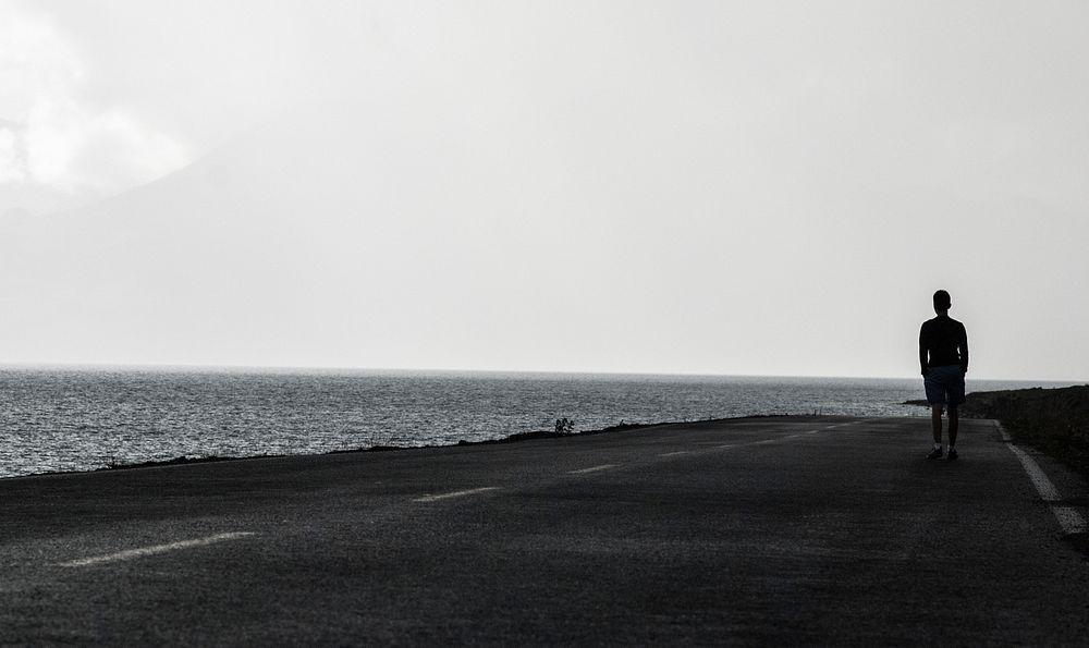 Black and white shot of lonely person walking on road near sea with clear sky. Original public domain image from Wikimedia…