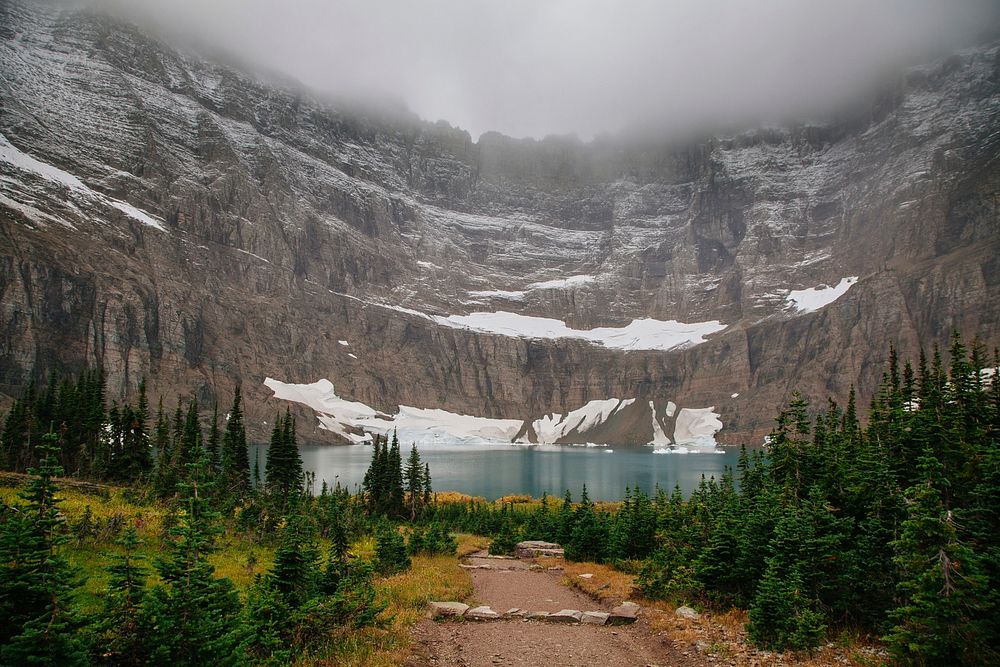 Fog circling above snowy mountains leading down to Iceberg lake surrounded by trees. Original public domain image from…