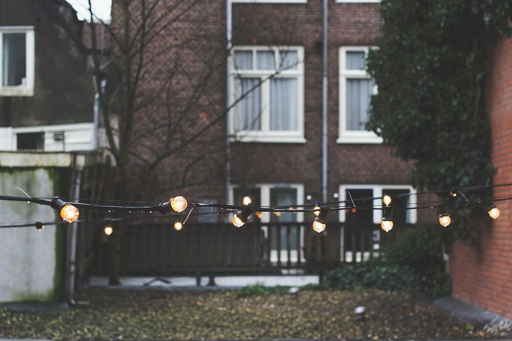 String lights in the back of an urban yard with a brick house and fences in the background. Original public domain image…