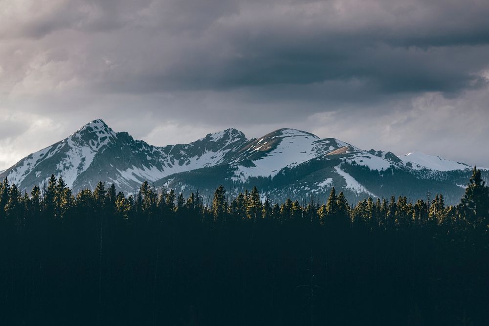 Dark gray clouds gathering over snow-capped mountain peaks behind a coniferous forest. Original public domain image from…
