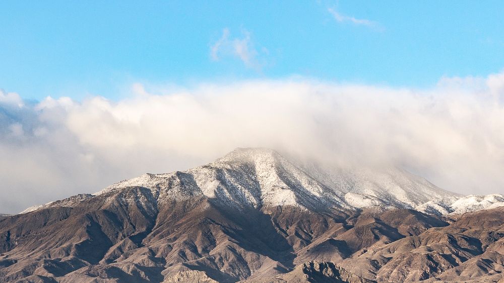 A scenic view of a snow covered mountain top with fog and a clear blue sky in Indio.. Original public domain image from…