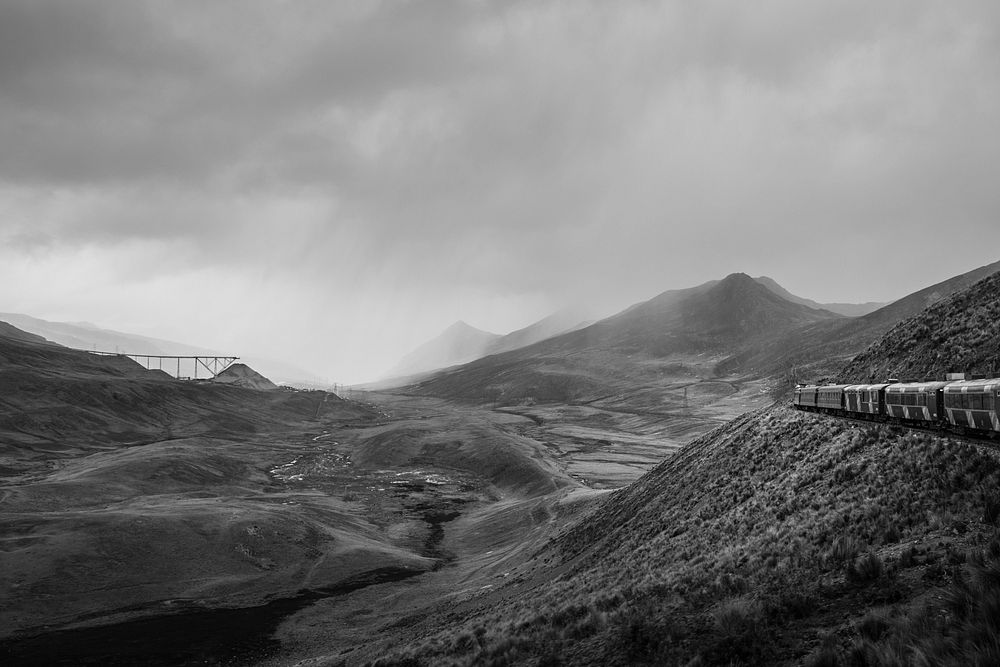Black and white panoramic shot of train travelling through mountains and rocks with cloudy sky. Original public domain image…