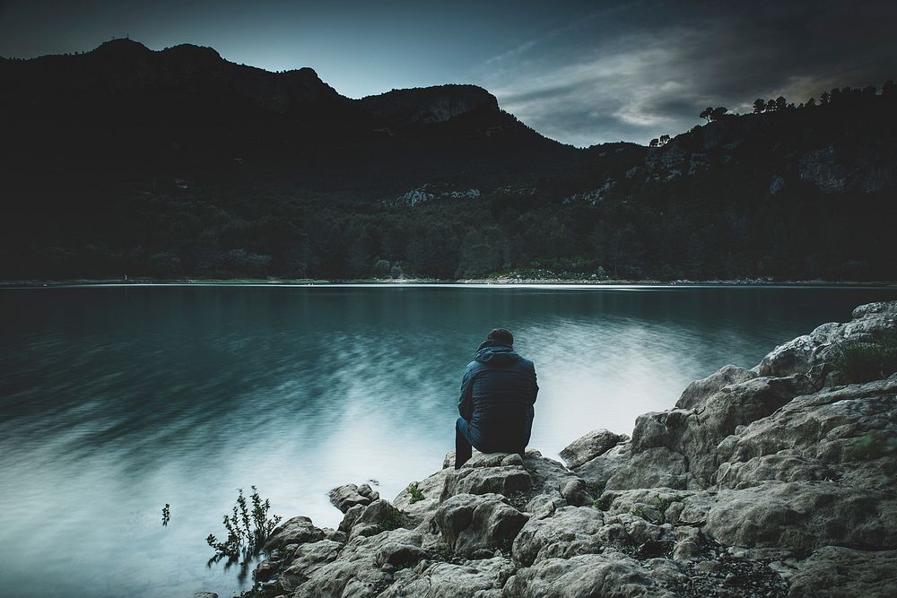 A man sitting on some rocks near a lake surrounded by mountains at dusk. Original public domain image from Wikimedia Commons