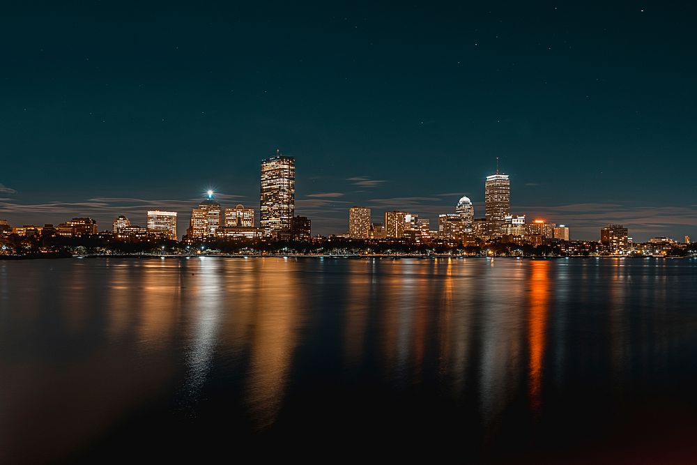 A bright skyline with a city waterfront seen from the water at night. Original public domain image from Wikimedia Commons