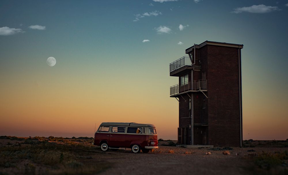 Campervan parked next to a standalone building in Dungeness a dawn-or-dusk with a faint moon in the orange-fading-to-blue…