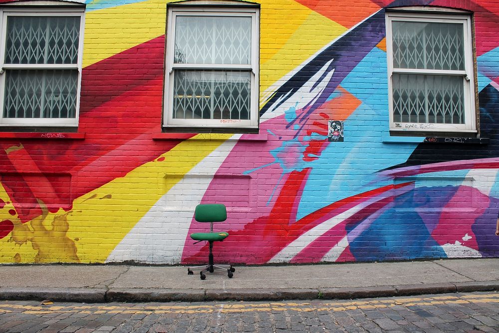 Green chair sits in front of colorful graffiti on brick building with three windows. Original public domain image from…