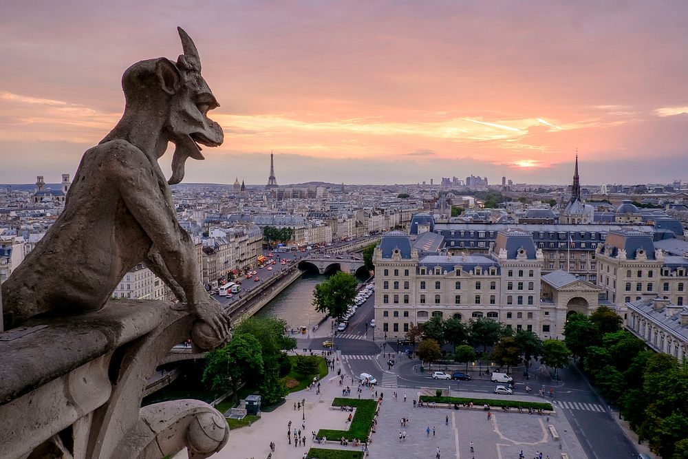 A gargoyle looks over the Notre-Dame Cathedral in Paris with a pale sunset lighting up the cityscape horizon. Original…