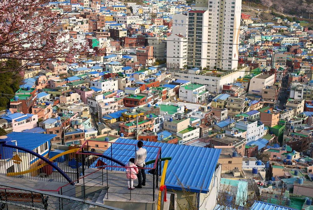 A father standing on a rooftop holding the hand of his child, looking out over the colorful rooftops of a city below.…