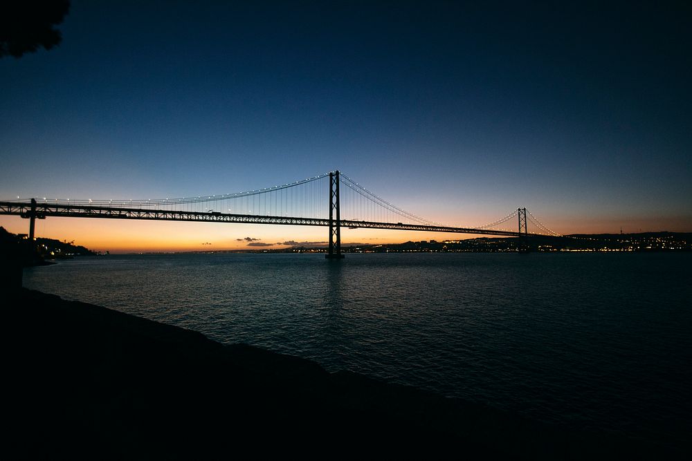 The end of a sunset behind a large suspension bridge over water in Lisbon.. Original public domain image from Wikimedia…