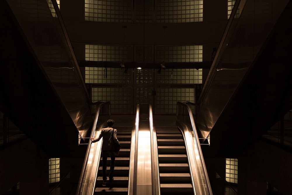 A shadowy shot of a man riding up a pair of escalators in the subway. Original public domain image from Wikimedia Commons