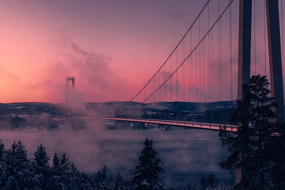 Clouds cover the Högakustenbron suspension bridge as a pink sunset covers the sky. Original public domain image from…