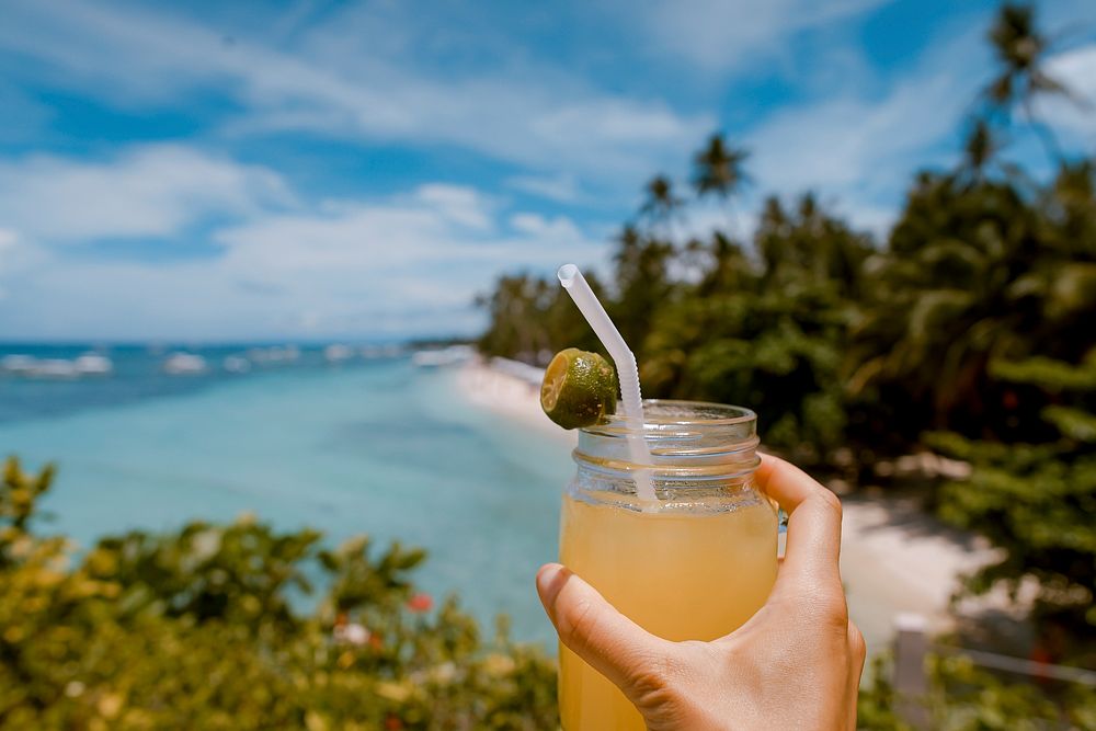 A person's hand holding up a cocktail with a lime and a straw against the tropical beach in the background. Original public…