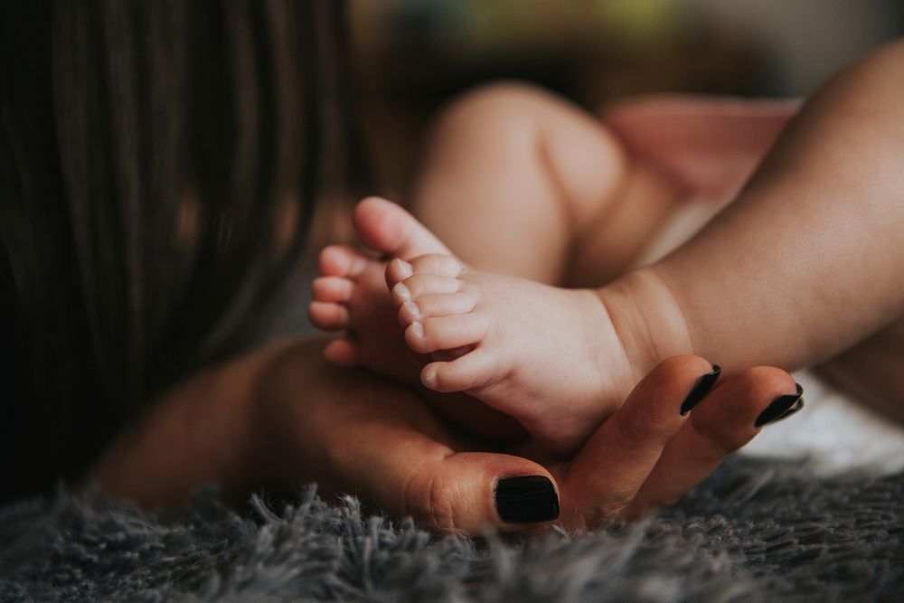 Infant's feet being held by a woman's hand with painted and manicured hands resting on a gray blanket. Original public…