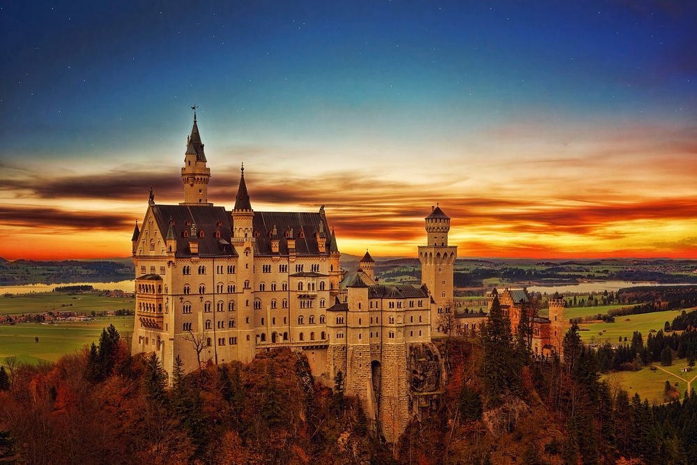 Drone view of Neuschwanstein Castle and country landscape with orange and blue sunrise-or-sunset horizon. Original public…