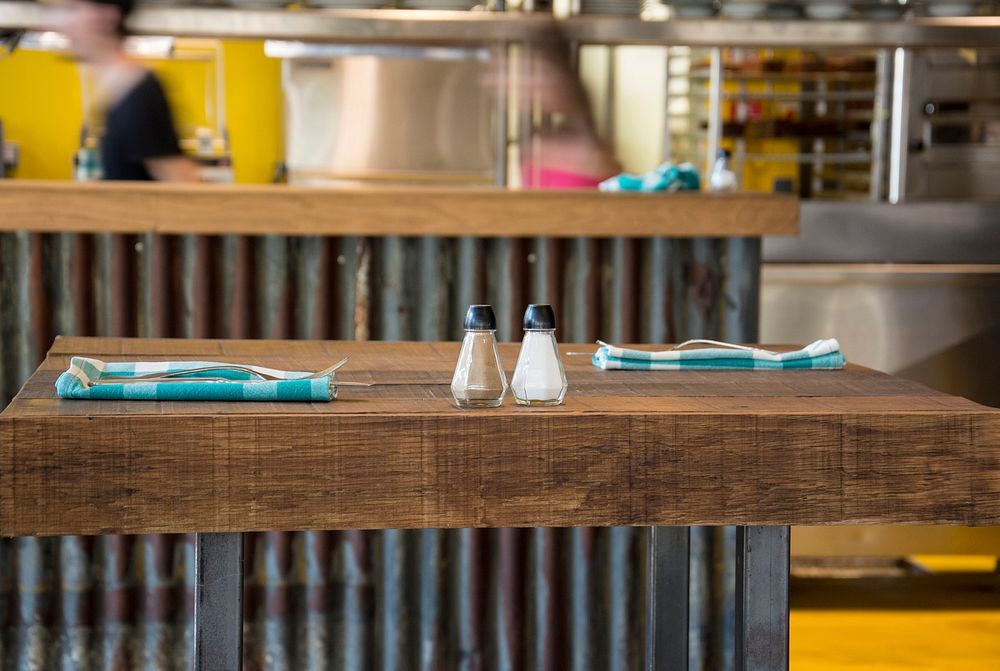 Salt and pepper shakers on a wooden table with blue plaid napkins and a busy kitchen in the background. Original public…