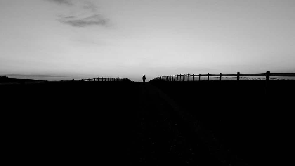 Black and white silhouette shot of person walking on horizon with wooden fence. Original public domain image from Wikimedia…