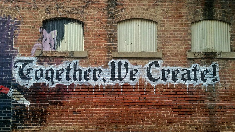 Graffiti words read "Together, we create!" below small windows on a brick wall. Original public domain image from Wikimedia…