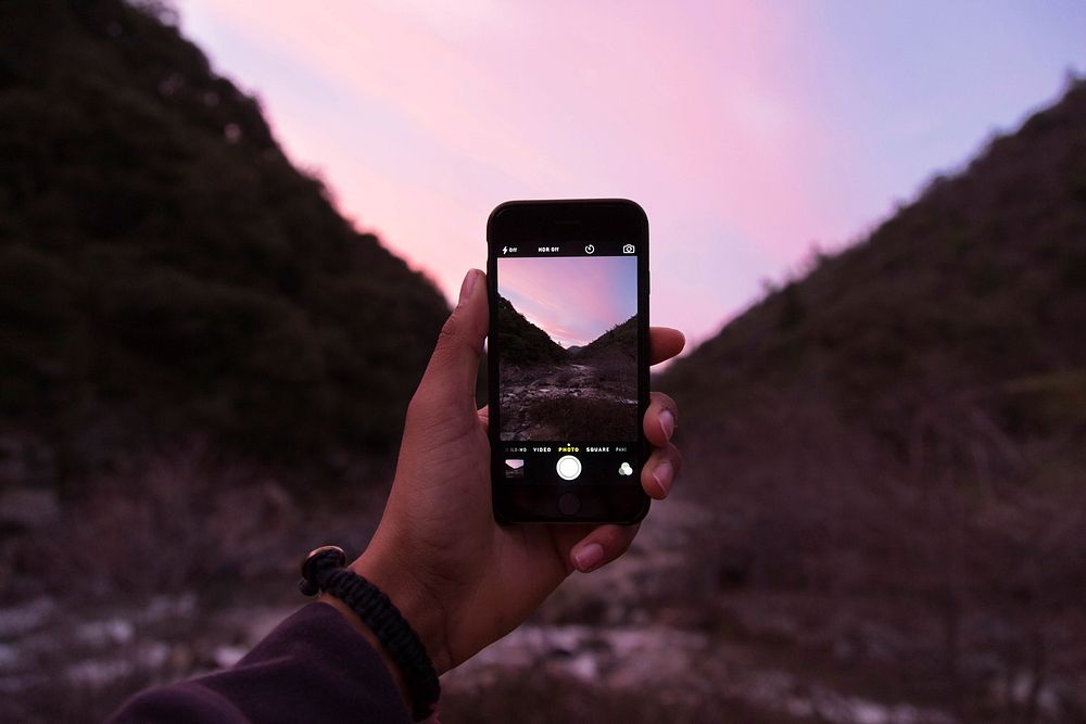 A person taking a photo of a pink sky and mountain with an iPhone Cellphone. Original public domain image from Wikimedia…