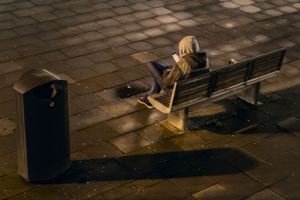 A hooded man on a bench using his smart phone beside a garbage can in a tile floor outside at night. Original public domain…