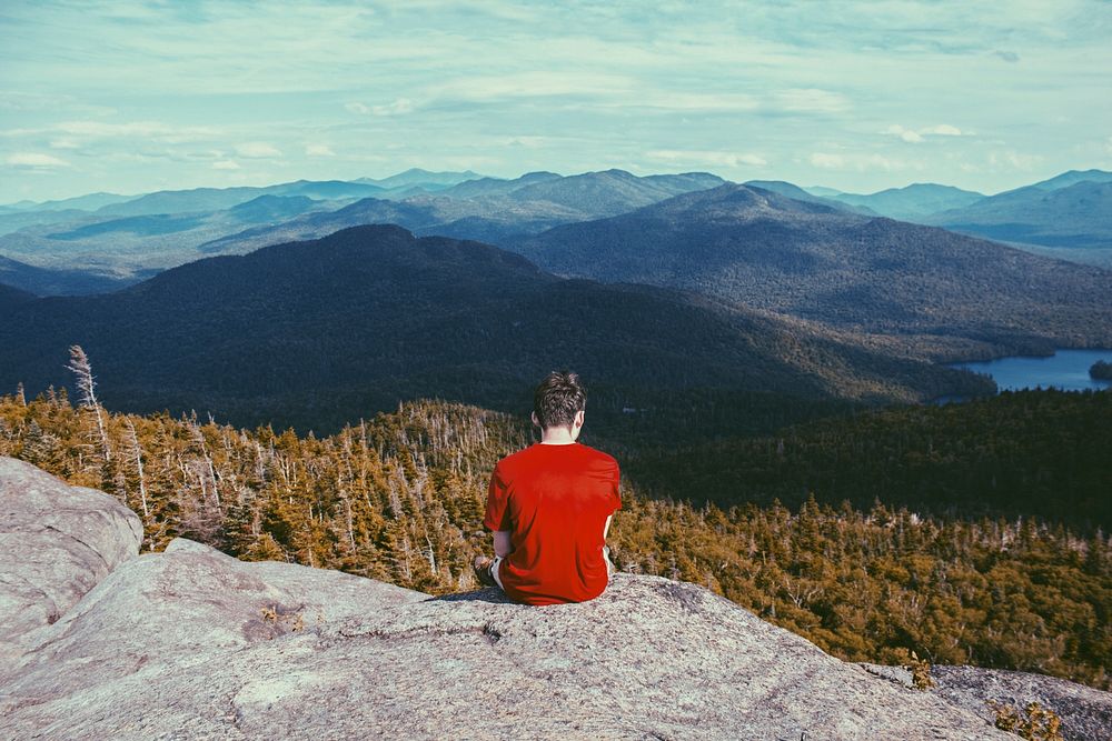 A man sitting on a rocky ledge overlooking wooded hills. Original public domain image from Wikimedia Commons