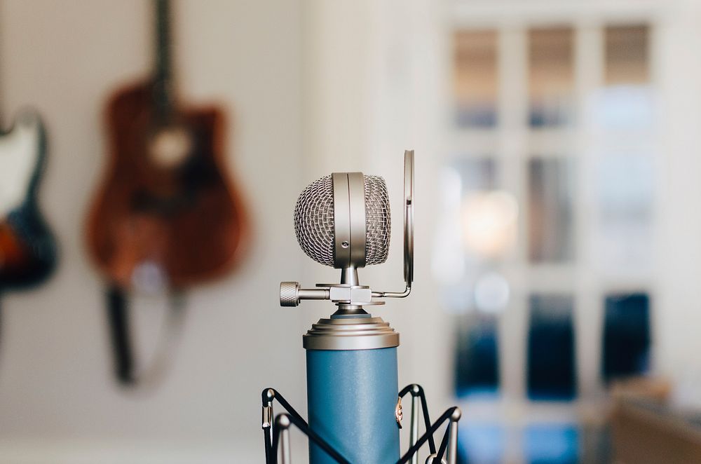 A small bluebird microphone on a stand with a pop filter in a white, bright room with hanging guitars. Original public…