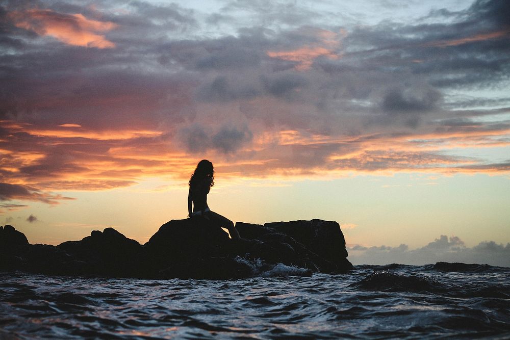 A sunset casts a silhouette of a Hawaiian mermaid sitting atop of rocks above the sea. Original public domain image from…