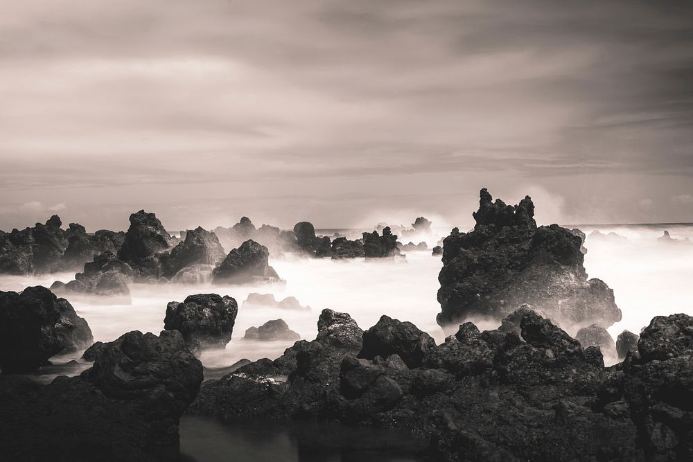 A grey image taken near Hana Highway, highlighting rocks in an ocean under a blanket of clouds. Original public domain image…