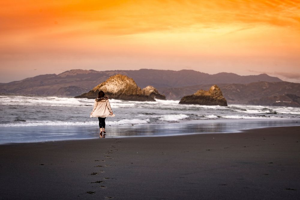 A woman walks across an empty beach as ocean waves crash on to the rocks and the sun sets in the sky. Original public domain…