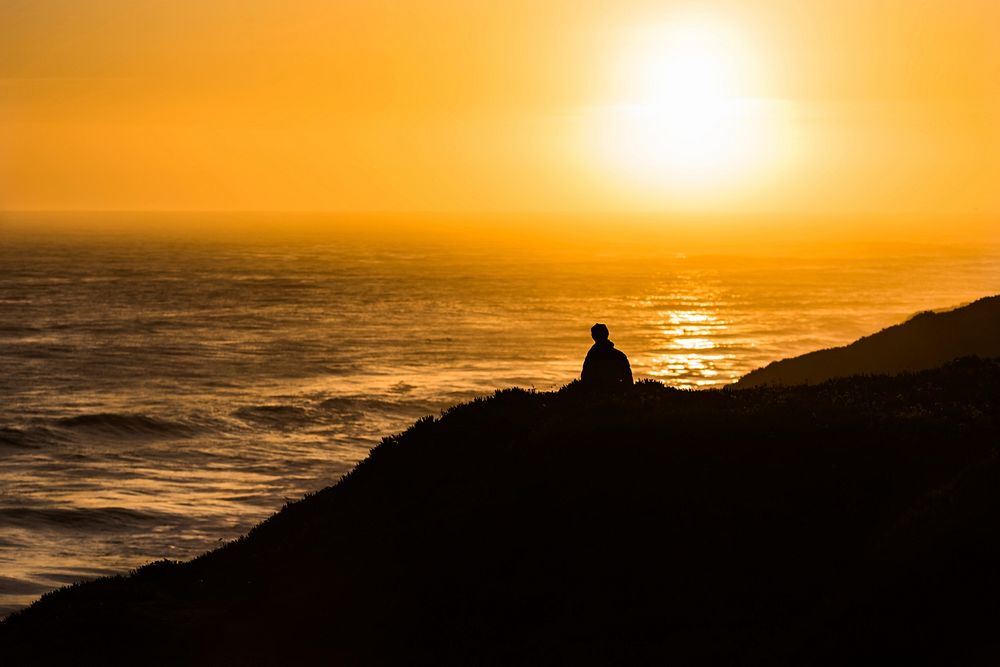 A lonely person sits atop a hill above the ocean of Santa Cruz, watching the orange sunset. Original public domain image…