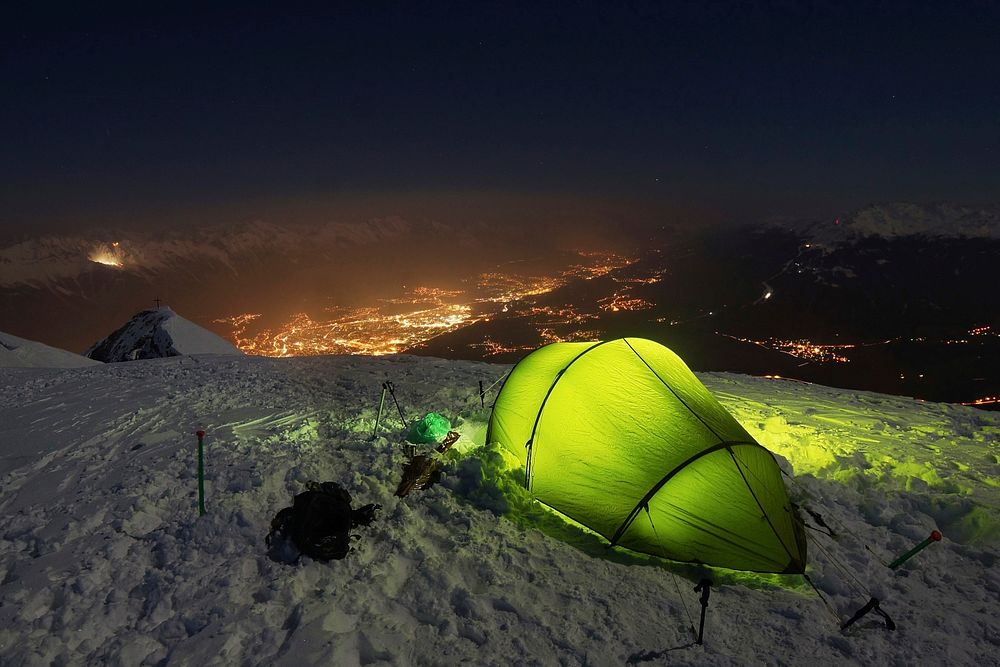 A green tent with green lights inside on top of a snow covered mountain overlooking the city lights.. Original public domain…
