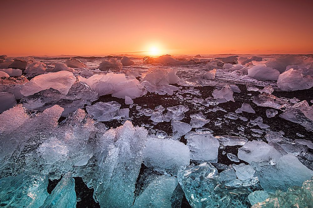 Ice breaks up on the shoreline at sunset against an orange sky and calm water in Iceland. Original public domain image from…