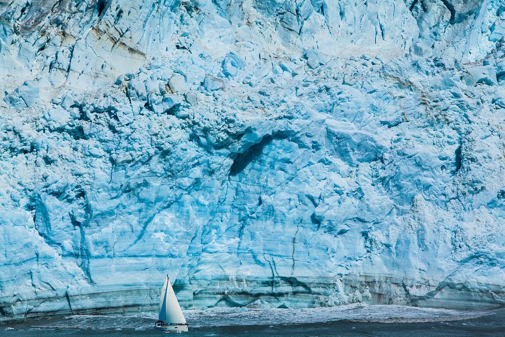 A steep light blue iceberg with a sailboat on the water below. Original public domain image from Wikimedia Commons