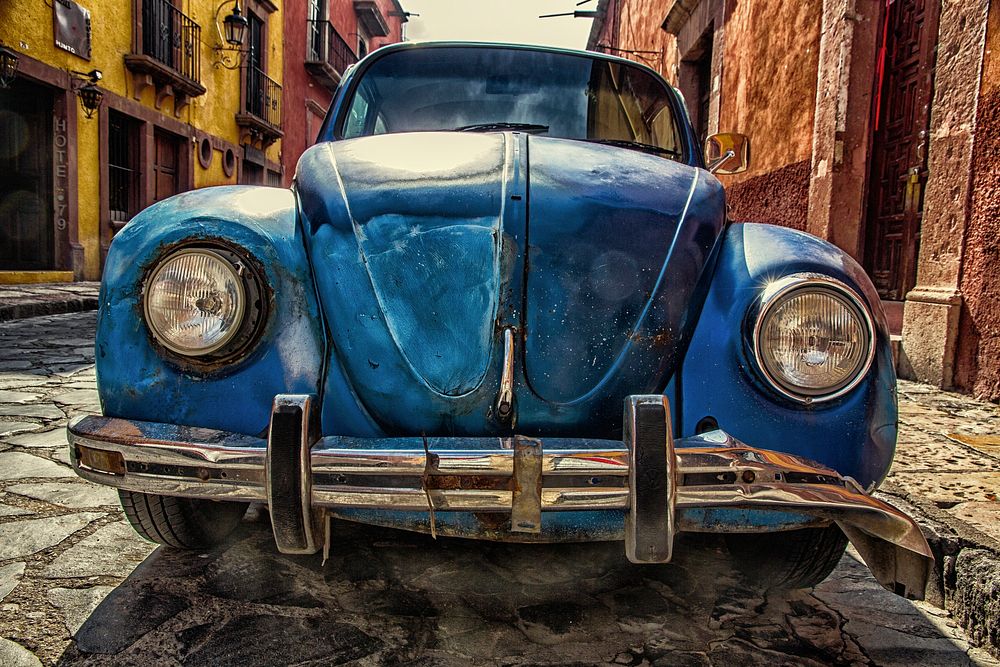 Front of a rusty, vintage, blue Volkswagen beetle on a cobblestone street, Blockbuster de San Miguel. Original public domain…