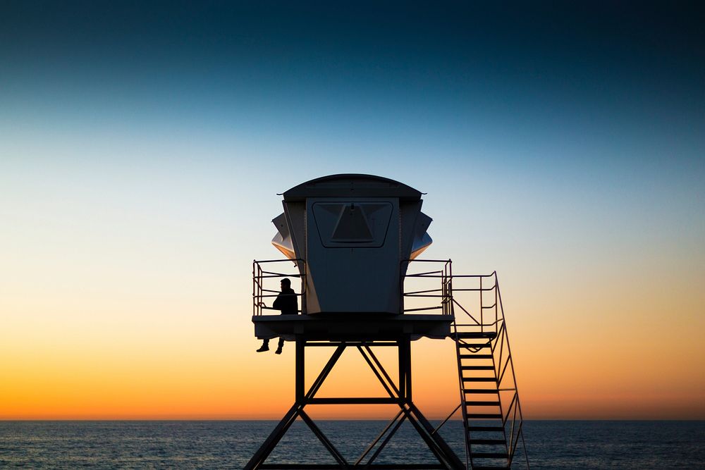 Silhouette of a man sitting alone against railing of a lifeguard tower, ocean and dusky sky in the background. Original…
