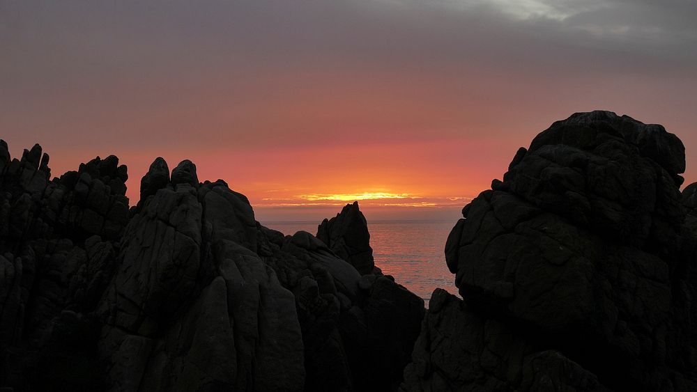 Large rocks and boulders by the sea, with view of the water and cloudy orange sunset between them. Original public domain…