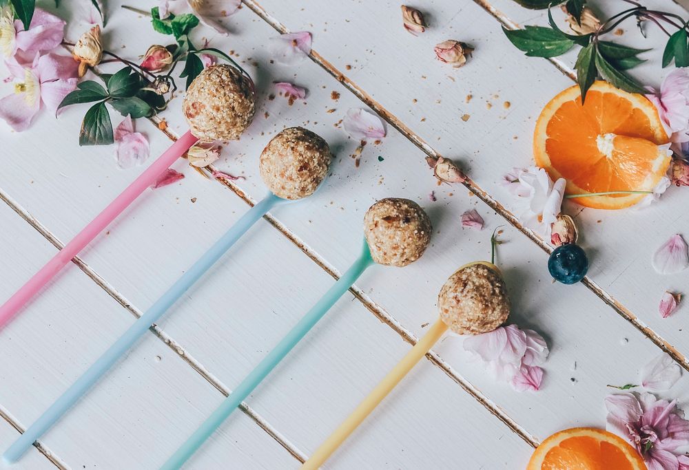 Rolled up balls of grain and oats on multicolored long plastic spoons beside sliced oranges, leaves, and flower petals on a…