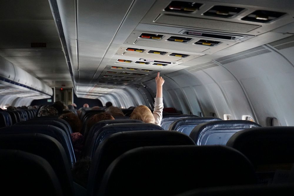 A child on a plane lifting their hand to adjust overhead controls above the seat. Original public domain image from…