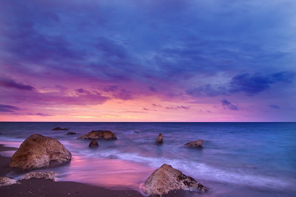 Rocks sit in a calm sea at twilight as the sunset creates a pink, purple, and navy sky at Playa del Peñón del Cuervo..…