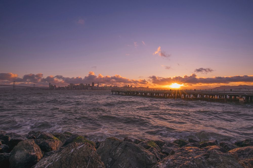 Sunset behind a pier with rough waves and gold light and a cityscape on the horizon.. Original public domain image from…