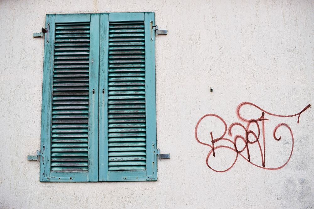 Red graffiti tag on white wall next to worn green window shutter. Original public domain image from Wikimedia Commons