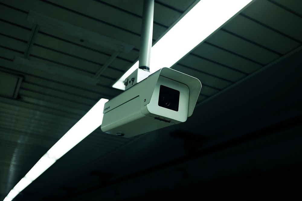 White surveillance camera monitoring a dark space with lined ceilings and overhead lights. Original public domain image from…
