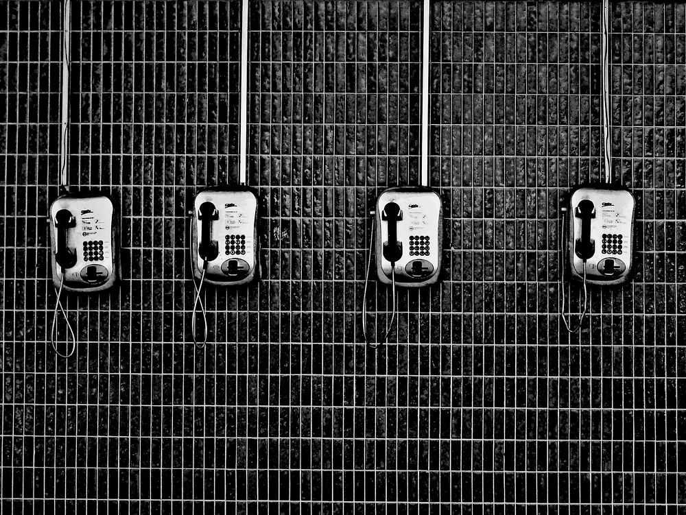 Black and white shot of four payphones on dark tiled wall. Original public domain image from Wikimedia Commons